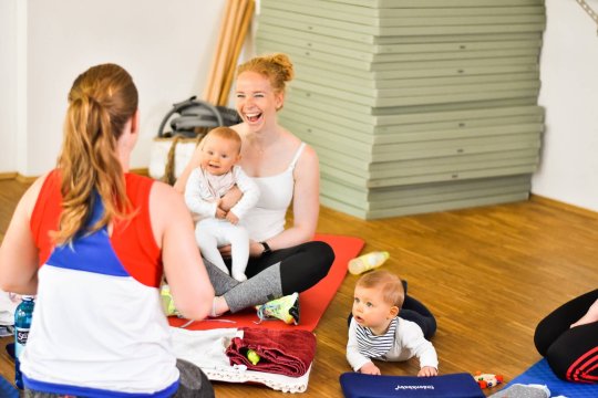 Eine lachende Frau sitzt mit zwei Kleinkindern in einer Turnhalle