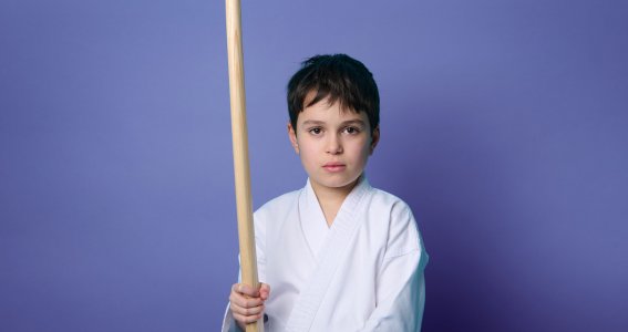 Ein Junge in Kampfsportkleidung mit einem Holzstock vor blauem Hintergrund